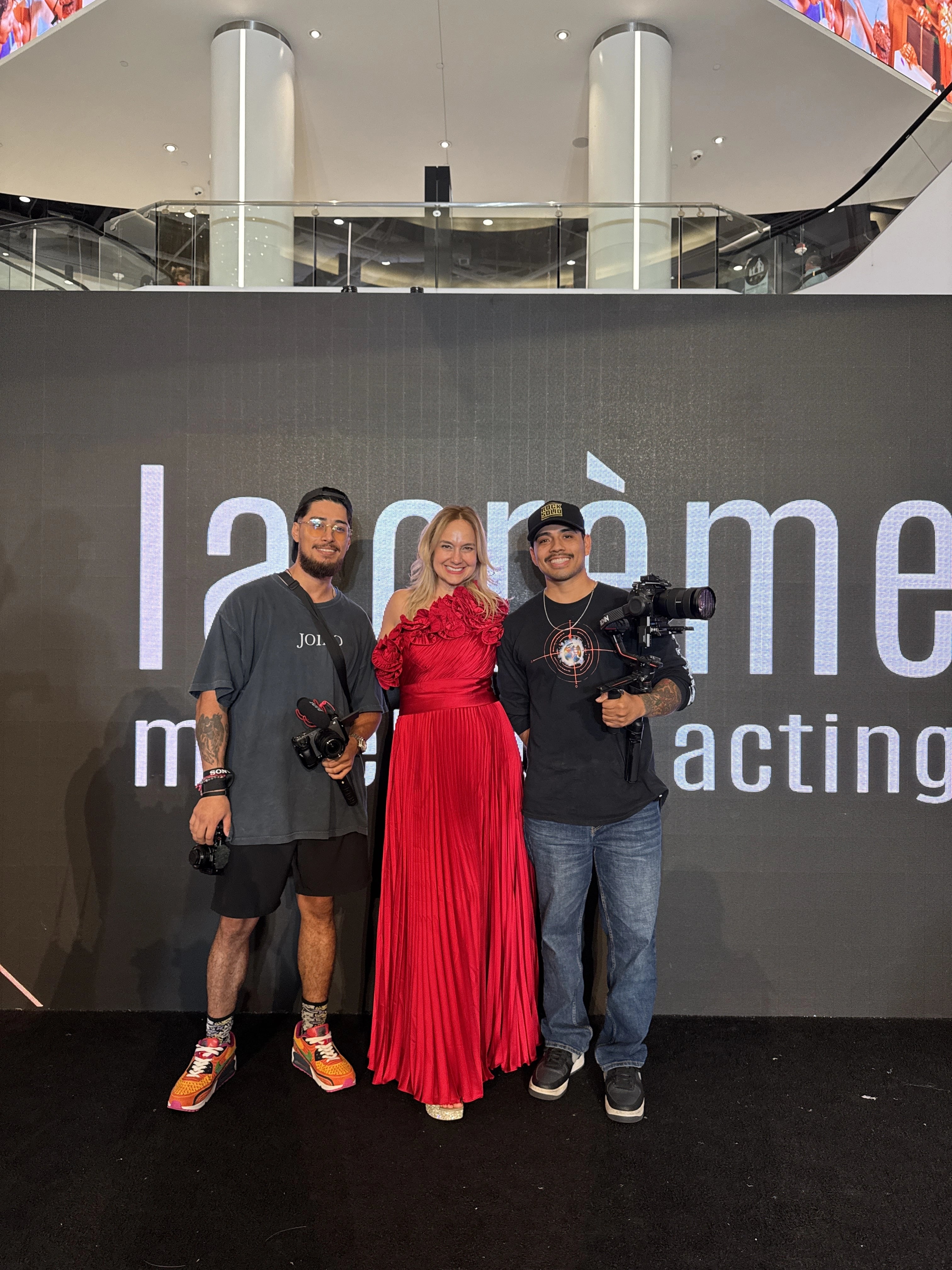 Haute Shots camera crew posing with a guest in a red gown in front of the LaCreme fashion show backdrop.